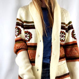 May include: Cream-colored cardigan sweater with a shawl collar and button closure. The sweater features brown, orange, and green geometric patterns on the sleeves and chest, with two front pockets. A vintage-inspired knitwear piece.