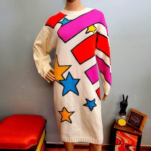 Könnte beinhalten: Cremefarbenes Strickkleid mit einem lebendigen Sternmuster in Rot, Pink, Gelb und Blau. Das Kleid hat einen Rundhalsausschnitt und lange Ärmel. Die Sterne sind schwarz umrandet.
