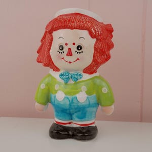 Può includere: Una statuetta di bambola in ceramica con capelli rossi, un cappello da marinaio bianco e un completo verde e blu. La bambola ha un viso dipinto con un papillon e scarpe nere. La statuetta è su una superficie bianca.