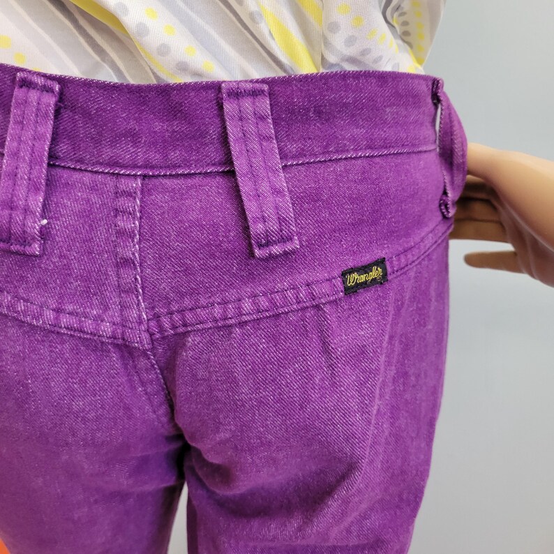 May include: Close-up of the back of purple Wrangler jeans. The jeans have belt loops, a back pocket, and a small black label with the Wrangler logo. The fabric appears to be denim.