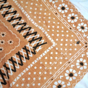 Op de afbeelding: Een beige en wit handdoek met een patroon van kleine bloemen, stippen en dikke zwarte zigzaglijnen. Het textiel heeft een zacht, getextureerd oppervlak met een duidelijke geometrische rand en bloemmotieven over de gehele stof.