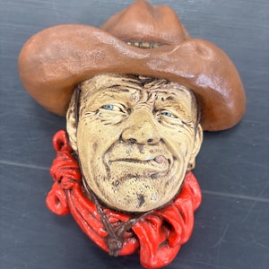 May include: A sculpted wall plaque featuring the head of a cowboy wearing a brown hat and a bright red neckerchief. The figure has a weathered, textured face with blue eyes and a small cigar held between the lips, rendered in a realistic style.