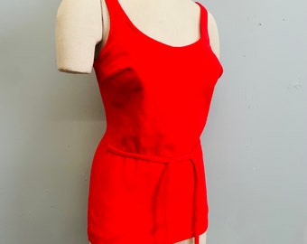 May include: A vibrant red tank top with a matching tie belt. The top features a scoop neckline and thin straps. The garment is displayed on a mannequin against a neutral gray background. The tank top is a solid color and appears to be made of a knit fabric.