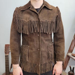 May include: Brown suede jacket with fringe detailing across the chest, shoulders, and pockets. The jacket has a button-up front and a collared neckline. A vintage-inspired piece.