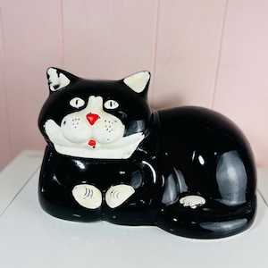 May include: A black and white ceramic cat figurine in a reclining position. The cat has a glossy black body, a white face, chest, and paws, with a bright red nose and mouth. The ears have white tips, and the eyes are white with black pupils.