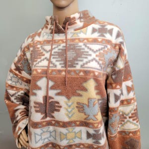 K&ouml;nnte beinhalten: Ein beigefarbener, brauner und blau gemusterter Fleece-Pullover mit hohem Kragen und Kordelzug. Das Design zeigt geometrische und vogelartige Formen. Das lang&auml;rmlige Oberteil wird auf einer Schaufensterpuppe pr&auml;sentiert.