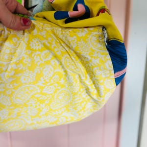 May include: A fabric bag with a yellow paisley lining. The exterior is made of various colourful fabrics, including a yellow background with a cartoon figure and blue details. The bag is held by a hand with red nail polish.
