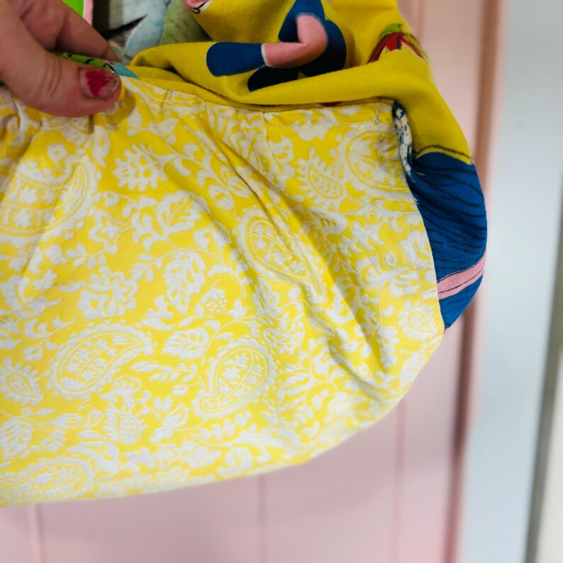 May include: A fabric bag with a yellow paisley lining. The exterior is made of various colourful fabrics, including a yellow background with a cartoon figure and blue details. The bag is held by a hand with red nail polish.