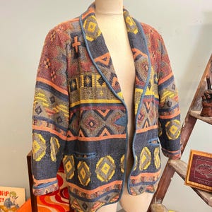 以下が含まれることがあります： ネイビー、オレンジ、イエロー、レッドの色合いの模様入りブレザー。幾何学模様のデザインで、青いトリムとシングルボタン留めが特徴です。ブレザーにはフロントポケットが2つあり、マネキンに飾られています。