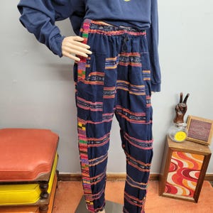 May include: Navy blue pants with colorful horizontal stripes and a drawstring waist. The pants have a relaxed fit and feature pockets. The stripes include shades of red, orange, yellow, and green. The pants are displayed on a mannequin.