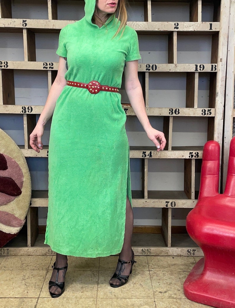 Puede incluir: Un vestido maxi verde con capucha, una abertura lateral y un cintur&oacute;n rojo con tachuelas. El vestido est&aacute; hecho de un tejido suave de toalla.