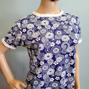 May include: A short-sleeved t-shirt featuring a dark blue background with a repeating white floral and spiral pattern. The garment has a solid white ribbed crew neckline and matching white ribbed trim on the edges of the short sleeves.