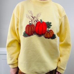 May include: A pale yellow fleece sweatshirt featuring a mock neck and long sleeves. The center of the chest displays an embroidered design of three pumpkins in shades of orange and red, accented with green leaves and tall stalks of wheat.