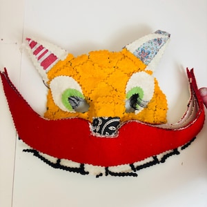 Puede incluir: Una máscara de zorro hecha a mano con una cara naranja brillante, ojos verdes y largas pestañas negras. La máscara presenta un gran bigote rojo y orejas blancas con detalles de tela estampada. La máscara está diseñada para disfraz o juego.