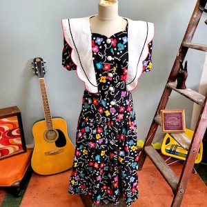 Può includere: Un abito nero vintage con un motivo floreale colorato e dettagli bianchi. L'abito presenta un colletto bianco smerlato con finiture rosse e bottoni bianchi. L'abito è esposto su un manichino.