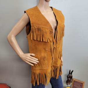 May include: A tan suede vest with a V-neck and fringed edges. The vest has two front pockets and a tie closure. The vest is sleeveless and has a vintage, bohemian style.