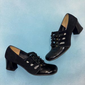 Puede incluir: Par de zapatos negros vintage con cordones, punta cuadrada y tacón de bloque. Los zapatos presentan una combinación de cuero liso y ante, con detalles recortados en los laterales. Los tacones miden unos 6,35 cm de alto.