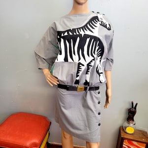 May include: Gray dress featuring a large black and white zebra design on the bodice. The dress has a black belt with a gold buckle and a skirt with black buttons. The sleeves are short, and the dress is displayed on a mannequin.