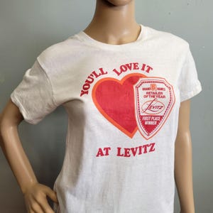 Puede incluir: Camiseta blanca con un corazón rojo y el texto "YOU'LL LOVE IT" en rojo. Debajo del corazón hay un escudo con las palabras "BRAND NAMES RETAILER OF THE YEAR" y "LEVITZ FIRST PLACE WINNER". El texto "AT LEVITZ" está debajo.