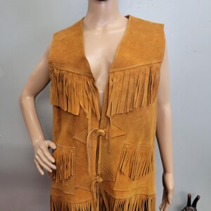 May include: A tan suede vest with fringe detailing. The vest has a V-neck, two front pockets, and fringe along the bottom and collar. The vest is sleeveless and has a tie closure at the front.