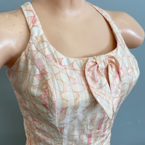 May include: Cream-colored halter dress with a floral pattern in pink and orange. The dress features a bow detail at the bust and a textured fabric. The dress is sleeveless and has a fitted bodice.