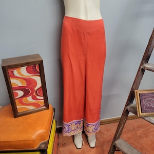 May include: Bright orange palazzo pants with a decorative border featuring purple and gold floral patterns. The pants are displayed on a mannequin, showcasing their wide-leg design. The pants are paired with white shoes.