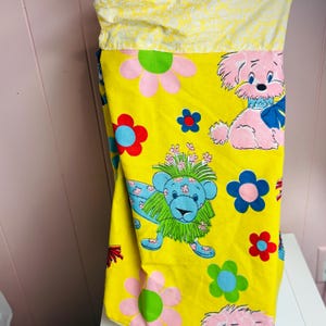 May include: A yellow pillowcase with a colourful cartoon print. The design features pink and blue cartoon animals, including a lion and a dog, and pink, blue, and green flowers. The top portion is a yellow fabric with a white floral pattern.