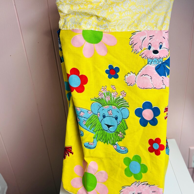 May include: A yellow pillowcase with a colourful cartoon print. The design features pink and blue cartoon animals, including a lion and a dog, and pink, blue, and green flowers. The top portion is a yellow fabric with a white floral pattern.