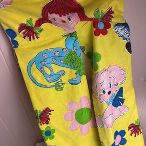 May include: Yellow fabric featuring a repeating pattern of cartoon characters, flowers, and bows. The characters include a girl with red plaits, a blue creature, and a pink dog. The fabric has a white lace trim.
