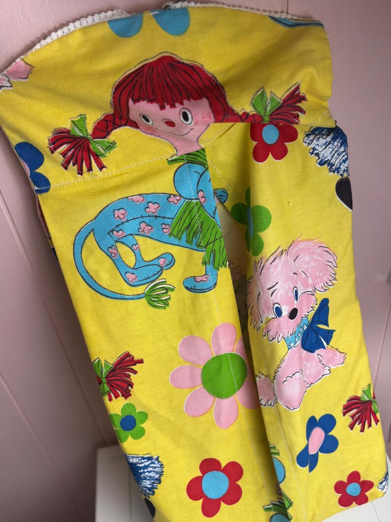 May include: Yellow fabric featuring a repeating pattern of cartoon characters, flowers, and bows. The characters include a girl with red plaits, a blue creature, and a pink dog. The fabric has a white lace trim.