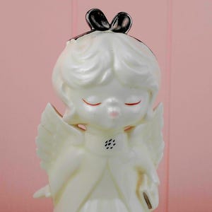 May include: A vintage ceramic angel figurine in ivory with closed eyes and a small pink mouth. The angel has white wings and a black bow on top of its head. The figurine is a decorative item.