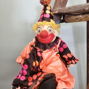 May include: A plush clown doll with a red nose, smiling mouth, and blue eyes. The clown wears a colorful outfit with a polka-dot pattern in shades of pink, orange, and brown. A matching hat with a pom-pom tops the head.