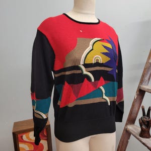May include: A vintage sweater with a colorful geometric design. The sweater features a red upper section, black sleeves and lower body, and a landscape-inspired pattern with triangles, clouds, and a sunburst in shades of red, blue, yellow, and brown.