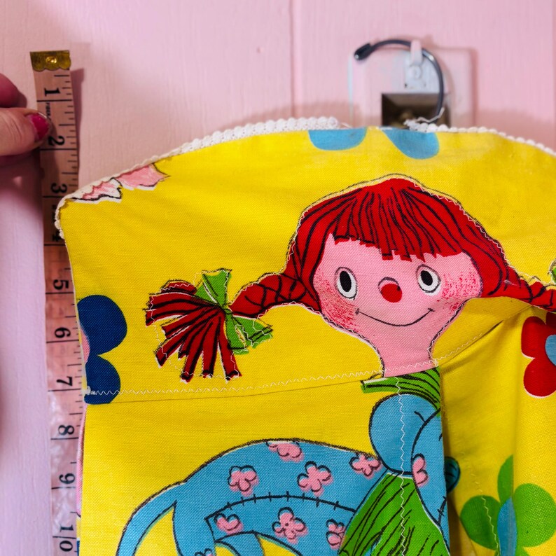 May include: A vintage yellow fabric doll with red braided hair, a pink face, and a blue dress. The doll is decorated with blue and red floral patterns. The doll is approximately 25 cm in length.