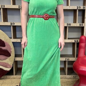 Puede incluir: Un vestido maxi de toalla verde con capucha y un cintur&oacute;n rojo con tachuelas. El vestido tiene mangas cortas y un corte holgado.