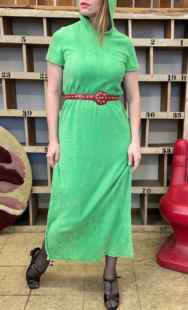 Puede incluir: Un vestido maxi de toalla verde con capucha y un cintur&oacute;n rojo con tachuelas. El vestido tiene mangas cortas y un corte holgado.