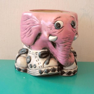 Puede incluir: Una maceta de cerámica rosa con forma de elefante, con una trompa, orejas y colmillos detallados. El elefante lleva un atuendo blanco y negro con lazos. La maceta tiene una amplia abertura para plantas.