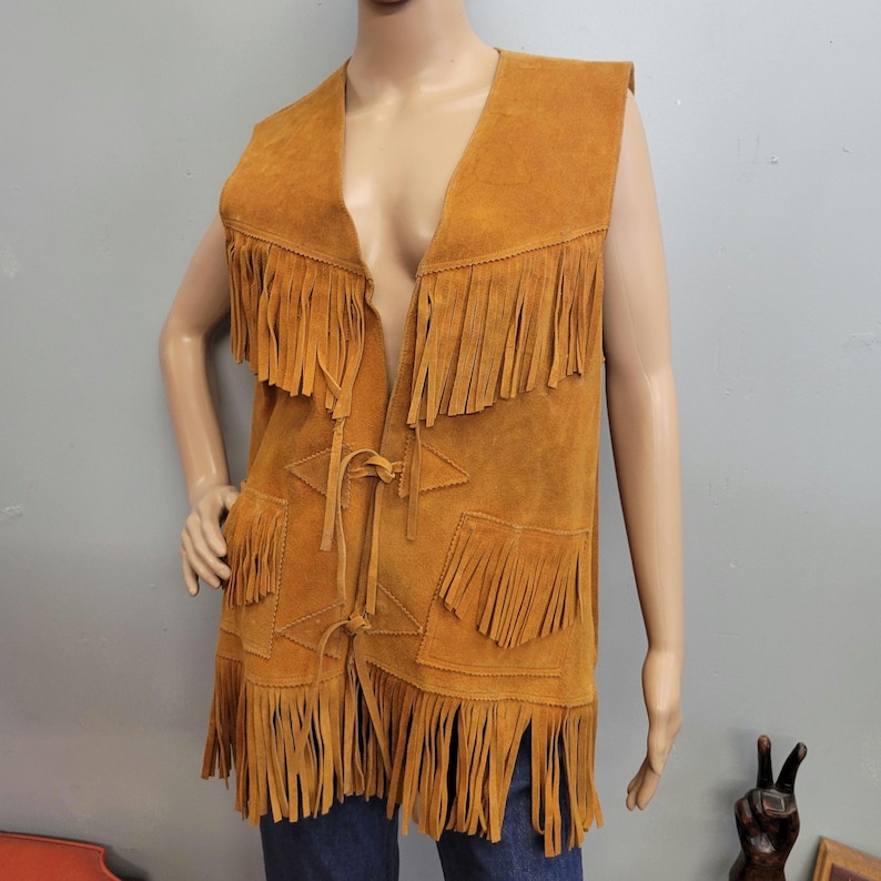 May include: A tan suede vest with fringe detailing. The vest features a V-neck, two front pockets, and a tie closure. The fringe is along the bottom, pockets, and shoulders. The vest is sleeveless and has a vintage, bohemian style.