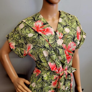 May include: A wrap dress featuring a vibrant floral pattern in shades of green, pink, and black. The dress has short sleeves, a V-neck, and a matching tie belt. The fabric has a textured appearance.