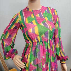 Może przedstawiać: Kolorowa sukienka w stylu vintage z geometrycznym wzorem. Sukienka ma długie rękawy i dopasowaną talię. Kolory obejmują róż, zieleń, żółty i fiolet. Lekki, zwiewny materiał nadaje jej retro charakter.