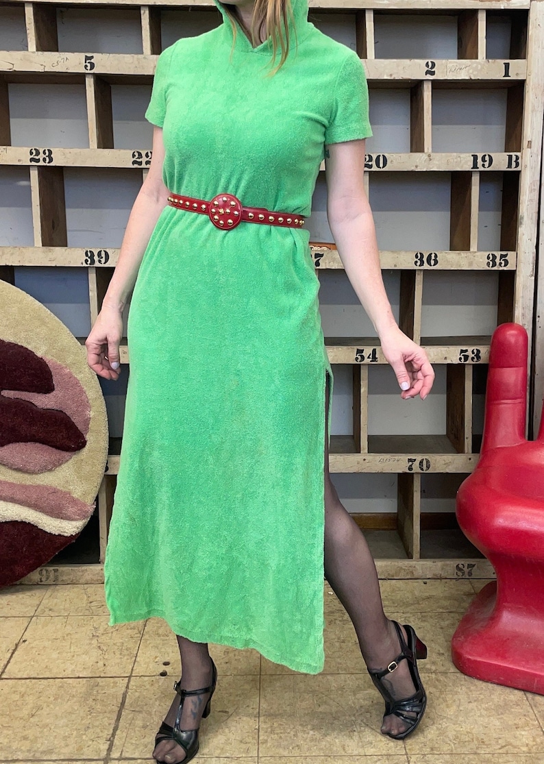 Puede incluir: Un vestido maxi de toalla verde con un cintur&oacute;n rojo tachonado. El vestido tiene cuello alto y mangas cortas. El vestido tiene una abertura lateral.