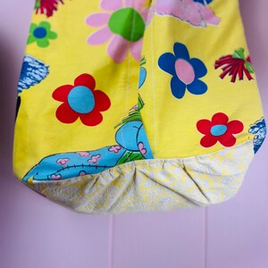 May include: A yellow tote bag with a floral pattern featuring red, blue, and pink flowers. The bag has a contrasting patterned bottom and a blue and white design. The bag is made of fabric.