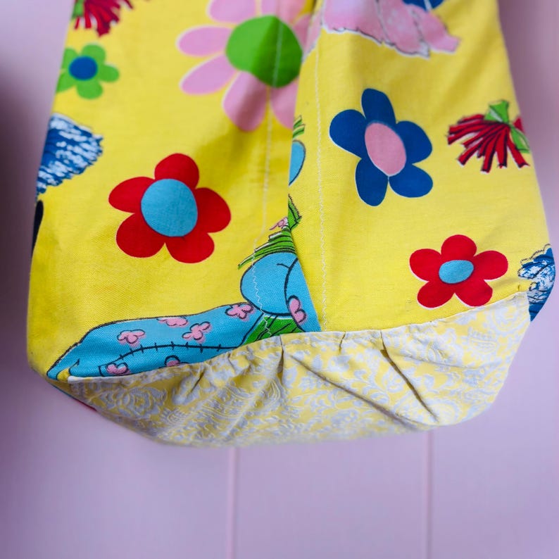 May include: A yellow tote bag with a floral pattern featuring red, blue, and pink flowers. The bag has a contrasting patterned bottom and a blue and white design. The bag is made of fabric.