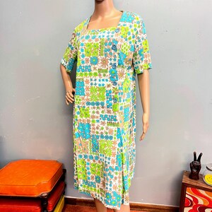 May include: A knee-length dress featuring a vibrant floral pattern in shades of bright green, turquoise, and tan on a white background. The garment has short sleeves, a square neckline, and a vertical row of five blue buttons along the front side.