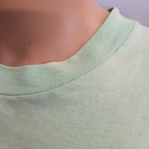 May include: Close-up of a light green t-shirt neckline. The collar is a slightly darker shade of green than the body of the shirt. The fabric appears to be a soft, knit material.
