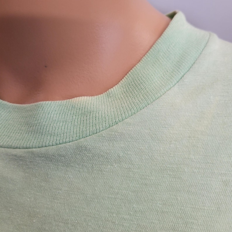 May include: Close-up of a light green t-shirt neckline. The collar is a slightly darker shade of green than the body of the shirt. The fabric appears to be a soft, knit material.