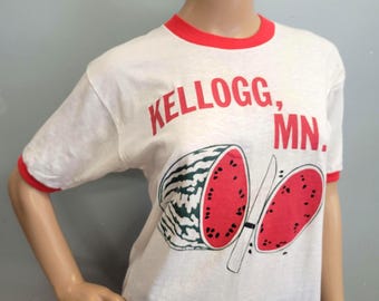 Może przedstawiać: Biała koszulka z czerwonymi wykończeniami z wzorem arbuza i napisem "KELLOGG, MN." Arbuz jest przekrojony na pół, z nożem pośrodku. Koszulka jest na manekinie.