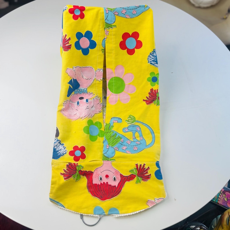 May include: A yellow fabric item featuring a playful design of cartoon animals, flowers, and foliage in red, pink, blue, and green. It has a scalloped edge and a metal ring at the bottom. The overall aesthetic is bright and cheerful.
