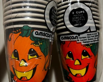Puede incluir: Dos pilas de vasos de papel con temática de Halloween. Cada vaso presenta un diseño de calabaza sonriente en naranja y rojo, con bordes a rayas blancas y negras. Los vasos están empaquetados en plástico transparente.