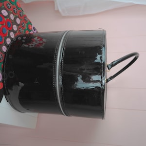 May include: A glossy black cylindrical handbag with a silver zipper running down the center. The bag has a curved handle and a shiny finish. The bag is a fashion accessory.